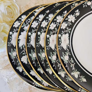 Puede incluir: Una pila de platos florales en blanco y negro con ribete dorado. Los platos presentan un diseño floral blanco sobre un fondo negro. Los platos tienen un borde dorado y un centro blanco.