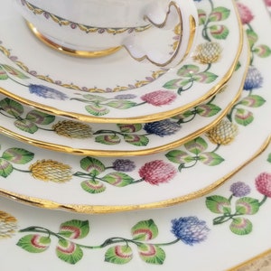 Może przedstawiać: Zestaw czterech białych porcelanowych filiżanek do herbaty i spodków ze złotą obwódką i kwiatowym wzorem. Wzór przedstawia zielone liście, różowe, niebieskie i żółte kwiaty.