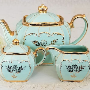 Peut inclure: Un service à thé vintage de couleur turquoise clair avec des bordures dorées. L'ensemble comprend une théière, un sucrier et un pot à crème. Chaque pièce présente un motif floral et des accents dorés, créant un look élégant et classique.