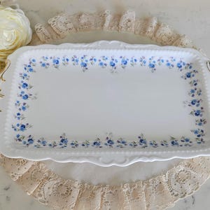Peut inclure: Un plateau de service rectangulaire blanc orné de motifs floraux bleus et de poignées dorées. Le plateau est posé sur un napperon en dentelle crème. Le design du plateau évoque la vaisselle vintage.