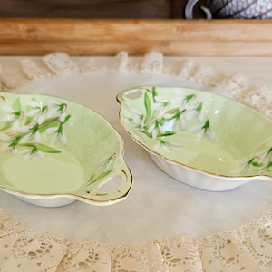 May include: Two oval-shaped, light green porcelain dishes with gold trim and handles. Each dish features a floral design of white flowers and green leaves. The dishes are placed on a white lace doily.