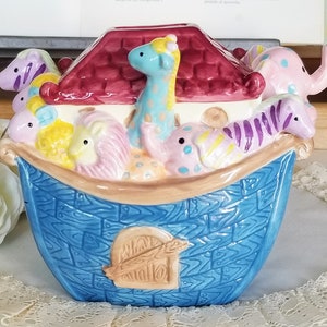 Op de afbeelding: Een keramische koektrommel in de vorm van een ark van Noach. De ark is blauw met een textuur van houtnerf. Het dak is bordeauxrood. De ark is gevuld met kleurrijke dierenfiguren, waaronder een giraf, een zebra en een olifant.