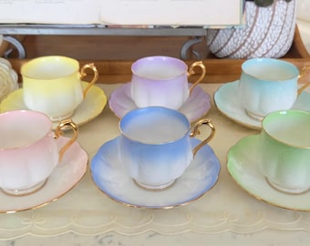 Puede incluir: Un juego de seis tazas de té y platillos de porcelana en colores pastel, incluyendo amarillo, lavanda, azul claro, rosa, azul medio y verde claro. Cada taza presenta un diseño acanalado, un asa con borde dorado y un platillo con borde dorado.