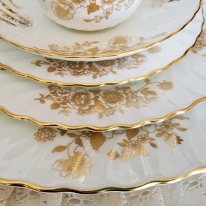 May include: A stack of white porcelain plates and a teacup featuring a scalloped edge with a gold trim. Each piece is decorated with a metallic gold floral pattern consisting of various blossoms and leaves arranged across the white surface.