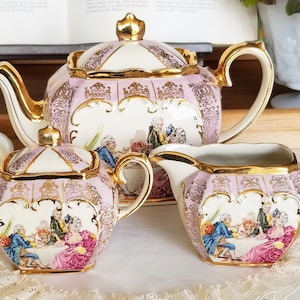 Könnte beinhalten: Ein rosa und goldenes Teeservice mit floralem Design. Die Teekanne, die Zuckerdose und die Milchkännchen sind alle mit einer Szene von Menschen verziert, die an einem Tisch sitzen. Das Set ist aus Porzellan und hat ein zartes, feminines Aussehen.