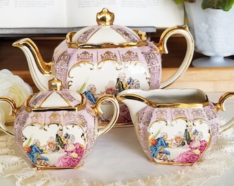 May include: A pink and gold tea set with a floral design. The teapot, sugar bowl, and creamer are all decorated with a scene of people sitting at a table. The set is made of porcelain and has a delicate, feminine look.