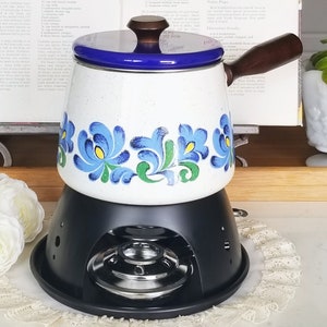 May include: White ceramic fondue pot with blue and green floral pattern, a black metal stand with a burner, and a wooden handle.