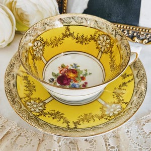 Puede incluir: Un juego de taza y platillo de té vintage. La taza y el platillo son de color amarillo brillante con filigranas doradas y detalles florales. El interior de la taza presenta un ramo de flores. El juego está colocado sobre un mantel de encaje blanco.