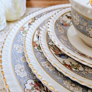 May include: A set of vintage porcelain tableware. The plates and teacup have a light blue floral and bird pattern with gold edging. The plates have a decorative, scalloped edge.