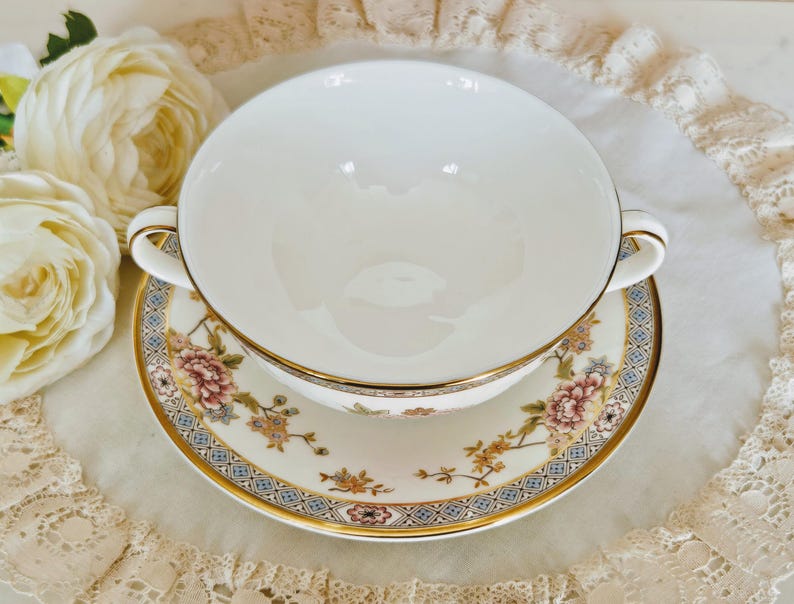 May include: A white porcelain soup bowl with handles, on a matching saucer. The saucer and bowl have a floral design with gold trim. The design includes pink, blue, and gold accents. A classic tea set.