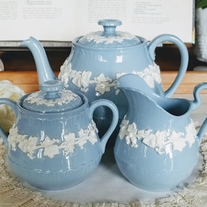 May include: A pale blue ceramic tea set featuring white floral relief patterns. The set comprises a teapot, a lidded sugar bowl, and a creamer. The items are displayed on a white lace doily, evoking a classic, vintage style.