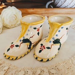 Peut inclure: Une paire de cache-pots en céramique en forme de bottines de couleur crème avec une finition brillante, une bordure jaune et des motifs floraux peints à la main avec des baies rouges et des feuilles vertes, des points bleus et une semelle rayée.