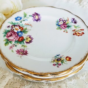 以下が含まれることがあります： 白い磁器の皿のスタック。縁はスカラップで、金色のトリムが付いています。各プレートは、ピンクのバラ、紫と青の花を特徴とするカラフルな花のブーケで飾られています。プレートは白いレースの表面に積み重ねられています。