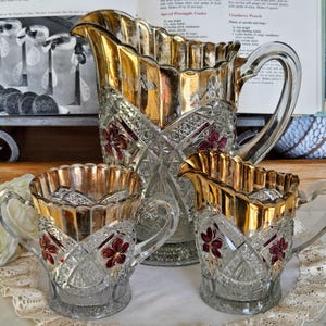May include: A set of three vintage glass jugs with gold accents and red floral designs. The jugs are made of clear glass and have a textured surface. The largest jug is a water jug, and the two smaller jugs are a creamer and a sugar jug.