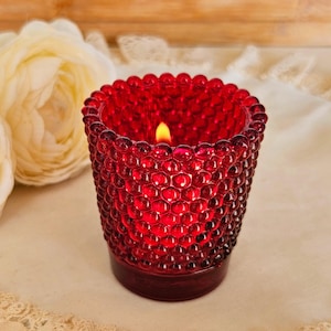 May include: A red glass votive candle holder with a textured, bubble-like surface. The candle holder is lit, with a small flame visible inside. The candle holder is sitting on a white surface with lace trim, and there are two white roses in the background.