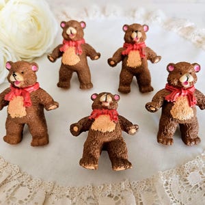 May include: Five brown bear figurines with red scarves and tan bellies are arranged on a white lace-trimmed surface. The bears have open mouths and pink accents on their ears and noses, giving them a cheerful appearance.