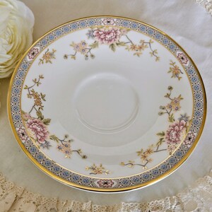 May include: A white porcelain saucer with a floral design featuring pink and yellow blossoms. The saucer has a gold rim and a blue and gold patterned border. The design is reminiscent of vintage china.