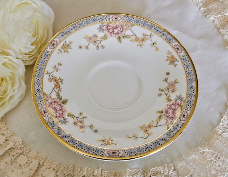 May include: A white porcelain saucer with a floral design featuring pink and yellow blossoms. The saucer has a gold rim and a blue and gold patterned border. The design is reminiscent of vintage china.