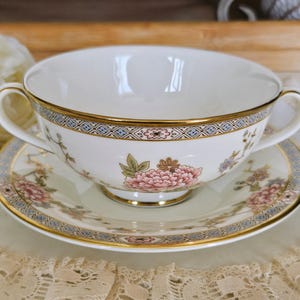 May include: A white porcelain soup bowl with two handles, and matching saucer. The set features a floral design with pink flowers and gold trim. The rim of the bowl and saucer have a blue and gold geometric pattern.