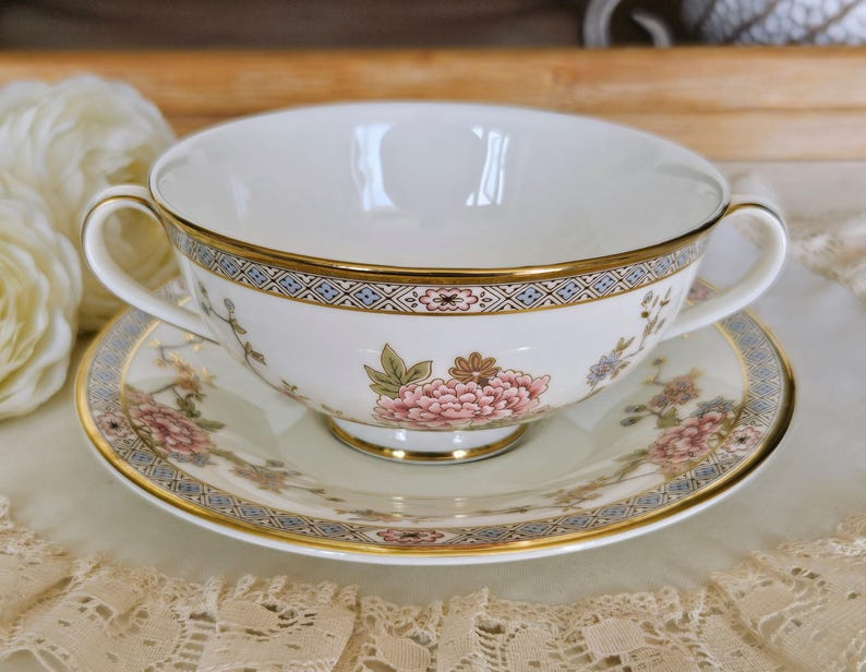 May include: A white porcelain soup bowl with two handles, and matching saucer. The set features a floral design with pink flowers and gold trim. The rim of the bowl and saucer have a blue and gold geometric pattern.