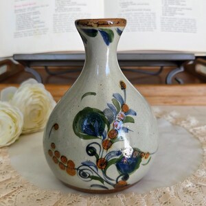 Peut inclure: Un vase en céramique artisanal avec un corps bulbeux et un col étroit. Le vase est peint avec des motifs floraux bleus, verts et oranges. Le bord est de couleur marron foncé. Le vase est posé sur un napperon en dentelle blanche.