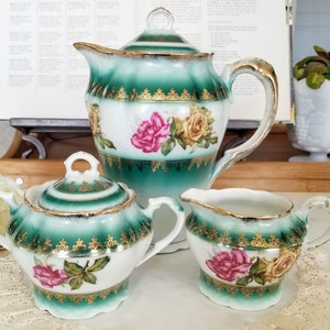May include: A porcelain tea set consisting of a teapot with a lid, a sugar bowl with a lid, and a creamer. Each piece features a white base with a deep green gradient, gold filigree trim, and painted pink and yellow rose floral motifs.