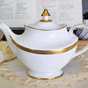 May include: A white porcelain teapot with gold detailing. The teapot features a curved spout, a rounded body, and a curved handle. The lid is topped with a gold finial. The teapot is displayed on a lace doily.
