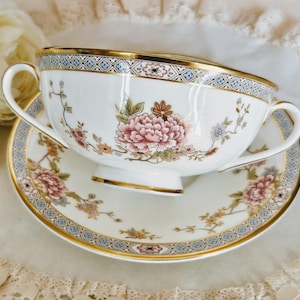 May include: A white teacup and saucer set with floral and gold detailing. The teacup has a large pink peony design and a gold rim. The saucer features a matching floral pattern and gold trim. The set is on a white surface.