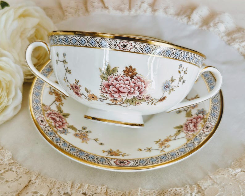 May include: A white teacup and saucer set with floral and gold detailing. The teacup has a large pink peony design and a gold rim. The saucer features a matching floral pattern and gold trim. The set is on a white surface.