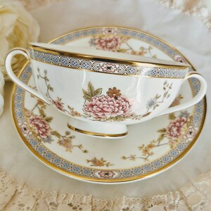 May include: A white teacup and saucer set with a floral design. The cup and saucer feature pink, peach, and blue flowers with gold trim. The rim of the cup and saucer has a blue and gold geometric pattern. The set is on a white surface.