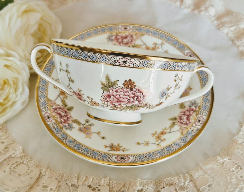 May include: A white teacup and saucer set with a floral design. The cup and saucer feature pink, peach, and blue flowers with gold trim. The rim of the cup and saucer has a blue and gold geometric pattern. The set is on a white surface.