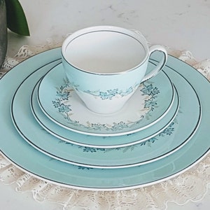 Puede incluir: Un juego de té vintage con un esquema de color azul claro y ribete negro. El juego incluye una taza de té, un platillo y dos platos, todos adornados con un patrón floral. El interior de la taza y el platillo es blanco. El juego está dispuesto sobre un tapete de encaje blanco.