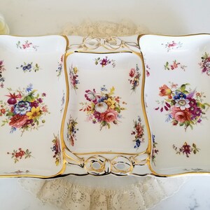 May include: A white porcelain serving dish with three compartments, decorated with colourful floral bouquets and gold trim. The dish has a bow-shaped accent in the centre, perfect for serving appetizers or desserts.