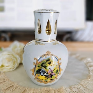 May include: White ceramic oil lamp with a gold-trimmed top and a decorative scene. The lamp features a rounded body and a detailed illustration of people in a landscape. The top has teardrop-shaped cutouts.