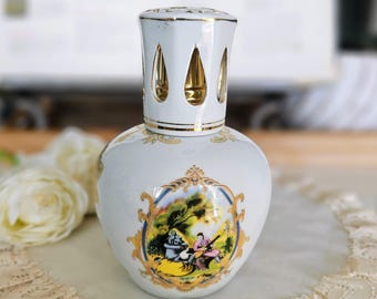 Könnte beinhalten: Weiße Keramik-Öllampe mit goldverziertem Deckel und dekorativer Szene. Die Lampe hat einen abgerundeten Körper und eine detaillierte Illustration von Menschen in einer Landschaft. Der Deckel hat tropfenförmige Aussparungen.