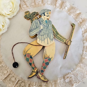 May include: A vintage wooden pull toy of a golfer with a plaid jacket and a golf club. The toy is painted in shades of brown, green, and red. The golfer is wearing a hat and has a mustache.
