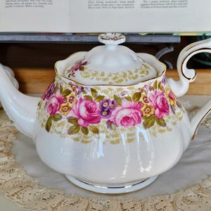 Puede incluir: Tetera de porcelana blanca con un diseño floral con rosas rosas y moradas, flores amarillas y hojas verdes. La tetera tiene un ribete dorado en el asa, el pico y la base. La tapa también está decorada con flores.