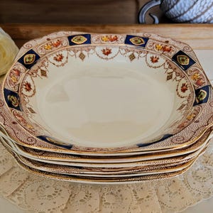 May include: A stack of four square ceramic bowls with rounded corners, featuring a cream-coloured centre and a decorative border of navy blue panels, gold filigree, and floral motifs in shades of orange, yellow, and green with gold trim.