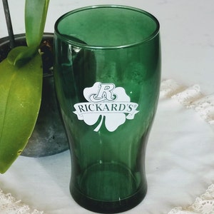 May include: A green pint glass with the Rickard's logo, featuring a shamrock and the brand name in white. The glass is empty and sits on a white lace-trimmed surface, with a plant in the background.