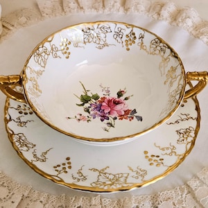 Könnte beinhalten: Weißes Porzellan-Suppenschüssel- und Untertassen-Set mit Goldrand und floralen Akzenten. Die Schüssel hat zwei goldene Griffe und ein Blumendesign in der Mitte. Die Untertasse hat einen passenden Goldrand und ein Blumendesign. Das Set ist im Vintage-Stil.
