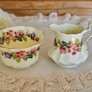 Puede incluir: Juego de azucarero y jarra de crema de cerámica color crema. Ambas piezas están adornadas con un estampado floral con flores rosas, moradas y verdes. La jarra de crema tiene un asa y ribete dorado, el azucarero tiene borde festoneado.
