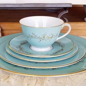 May include: A vintage tea set in light blue with gold trim and floral patterns. The set includes a teacup, saucer, and plates. The teacup, saucer, and plates have a gold floral design. The plates and saucer are stacked, with the teacup on top.
