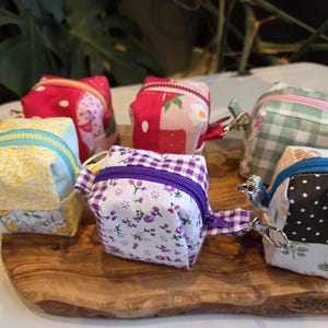 May include: A collection of small, square fabric pouches with zippers and key rings. The pouches feature various patterns, including floral, polka dots, and gingham, in colors like red, yellow, and green. They are arranged on a wooden board.
