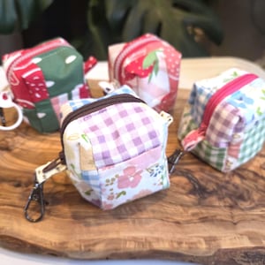 May include: Four small, square fabric pouches with zippers and keychains sit on a wooden board. The pouches feature various patterns, including gingham, floral, and fruit designs, in shades of red, green, pink, and blue.