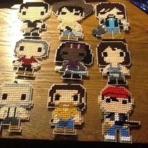 Puede incluir: Nueve personajes de punto de cruz de pixel art, cada uno con un dise&ntilde;o y una combinaci&oacute;n de colores diferente. Los personajes est&aacute;n todos en estilo chibi, con cabezas grandes y cuerpos peque&ntilde;os. Los personajes llevan todos atuendos diferentes, incluyendo una camisa roja, una camisa azul, una camisa amarilla, una camisa verde, una camisa morada, una camisa negra, una camisa blanca, una camisa marr&oacute;n y un sombrero rojo.