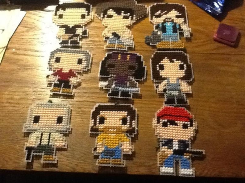 Puede incluir: Nueve personajes de punto de cruz de pixel art, cada uno con un dise&ntilde;o y una combinaci&oacute;n de colores diferente. Los personajes est&aacute;n todos en estilo chibi, con cabezas grandes y cuerpos peque&ntilde;os. Los personajes llevan todos atuendos diferentes, incluyendo una camisa roja, una camisa azul, una camisa amarilla, una camisa verde, una camisa morada, una camisa negra, una camisa blanca, una camisa marr&oacute;n y un sombrero rojo.