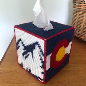May include: A blue, red, and white tissue box cover with a white mountain range and the state of Colorado's flag on the front.
