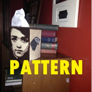 May include: A tissue box cover with a black and white portrait of a person with a dark background. The cover is decorated with a pattern of a wolf's head. The tissue box cover is sitting on a shelf with a stack of books behind it.