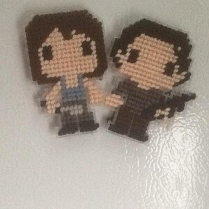 Puede incluir: Dos figuras de punto de cruz de pixel art, una con cabello casta&ntilde;o y una camisa azul, la otra con cabello casta&ntilde;o y una camisa negra.
