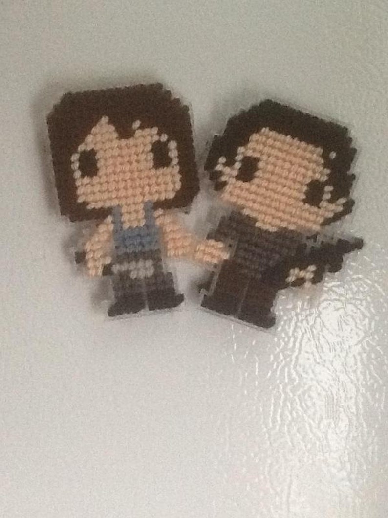 Puede incluir: Dos figuras de punto de cruz de pixel art, una con cabello casta&ntilde;o y una camisa azul, la otra con cabello casta&ntilde;o y una camisa negra.